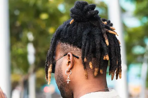 A man with styled black dreadlocks in a top bun, showcasing gold-tipped ends, wearing an earring with an ankh symbol, representing a trendy variation of MEN'S SHORT CURLY HAIRCUTS.