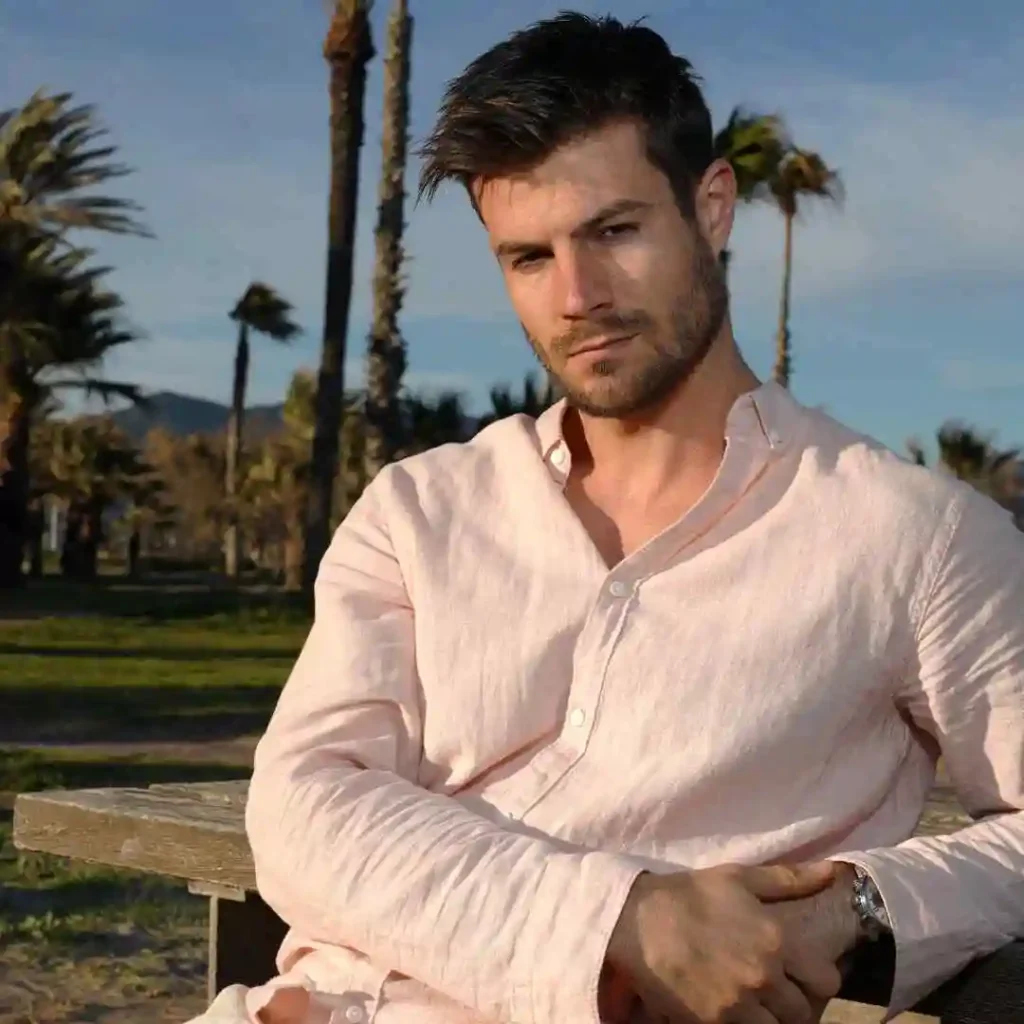 A man in a light pink shirt sits on a bench surrounded by palm trees against a clear blue sky, arms crossed with a relaxed demeanor.
