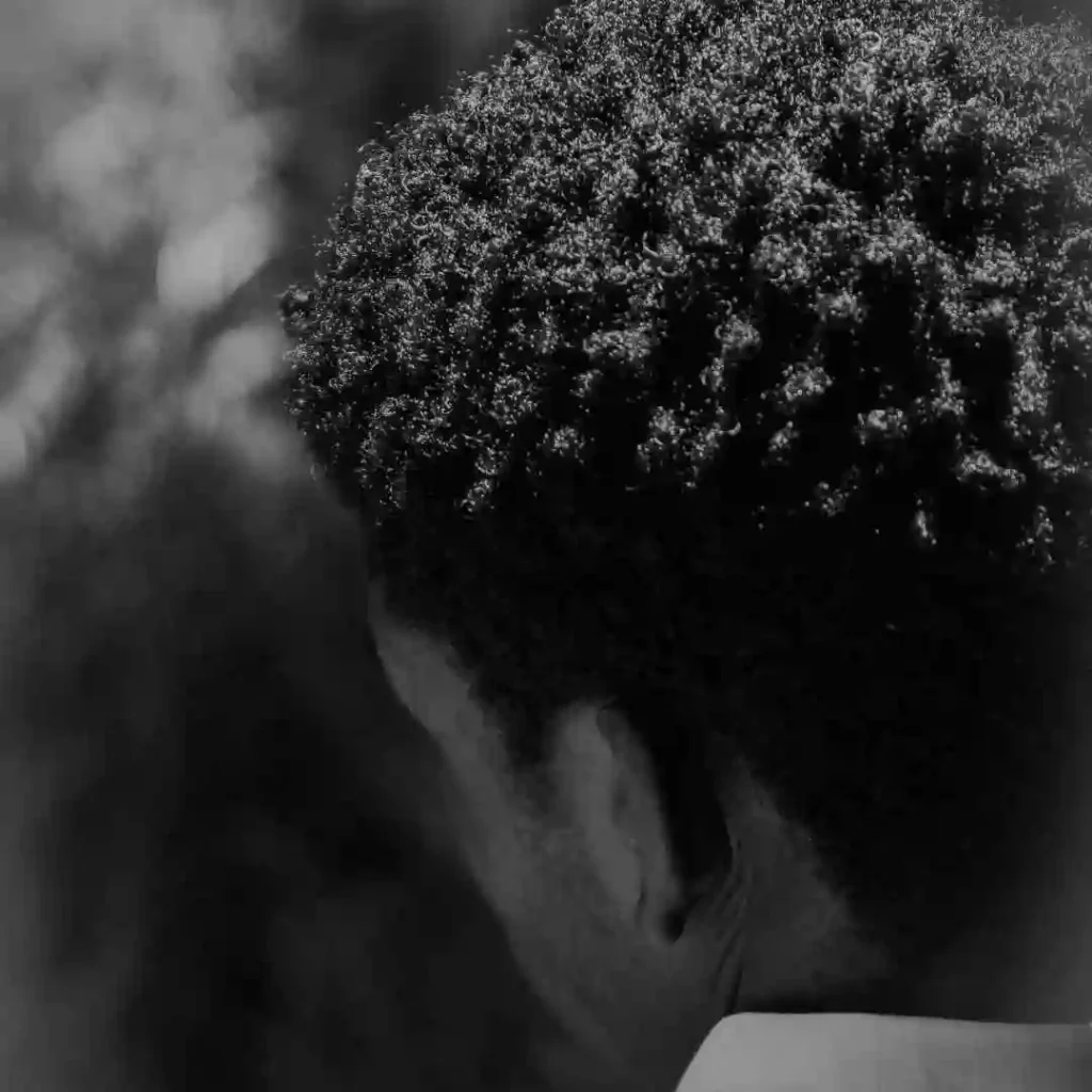 Close-up of a person’s head from behind, showcasing tight, textured curls and a softly blurred background. Black and white photograph.