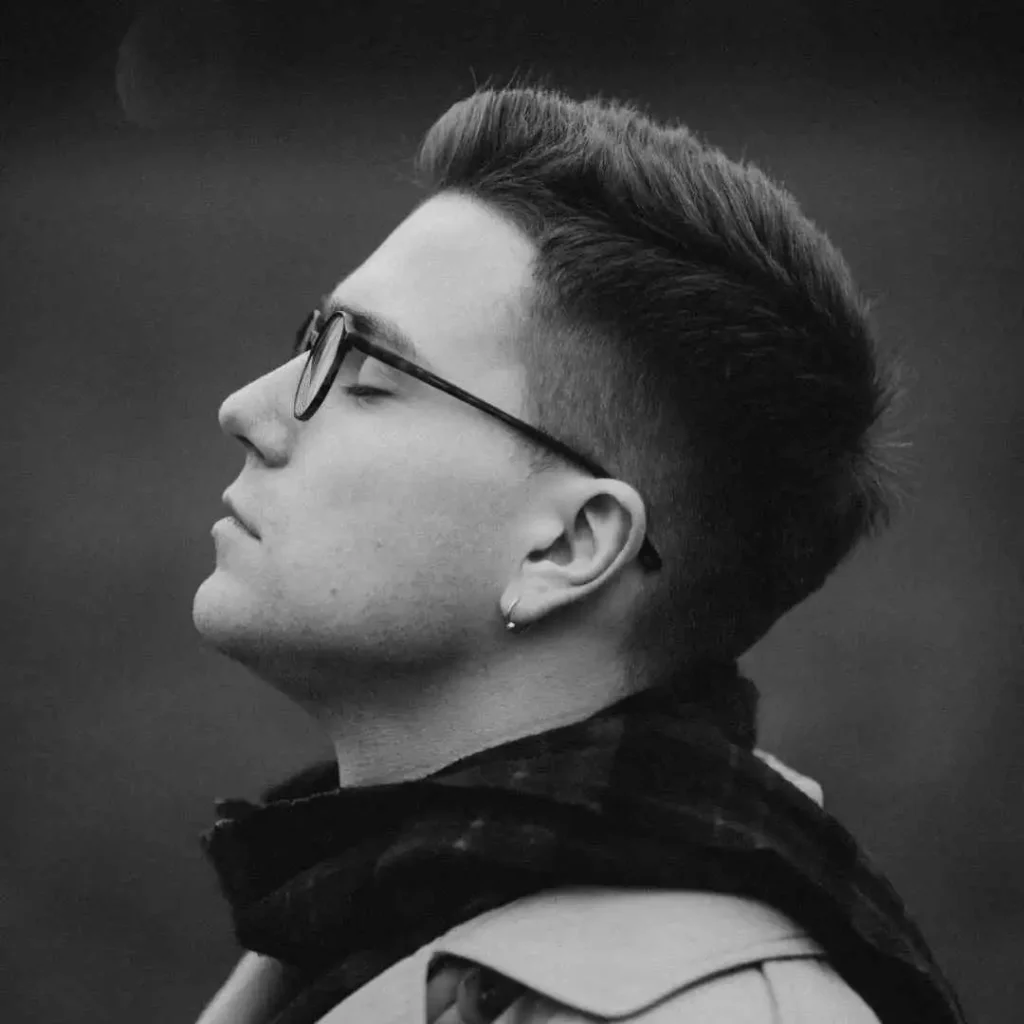 18 Top Men Hairstyles Quiff 3 "A person with a stylish men hairstyles quiff looks upward, wearing a light trench coat and a black scarf, set against a blurred dark background.