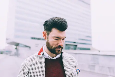 Discover 20 New Pompadour Hairstyles- A Comprehensive Guide 3 A stylish individual with POMPADOUR HAIRSTYLES, wearing a patterned jacket and sweater, stands outdoors in front of a modern building, exuding a contemporary vibe.