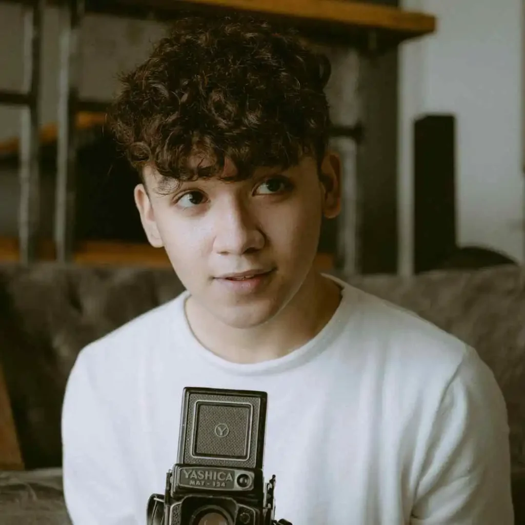 29 Best Men’s Short Curly Haircuts 3 A person with MEN'S SHORT CURLY HAIRCUTS wearing a white shirt holds a vintage Yashica camera, sitting on a plush couch in a cozy indoor setting.
