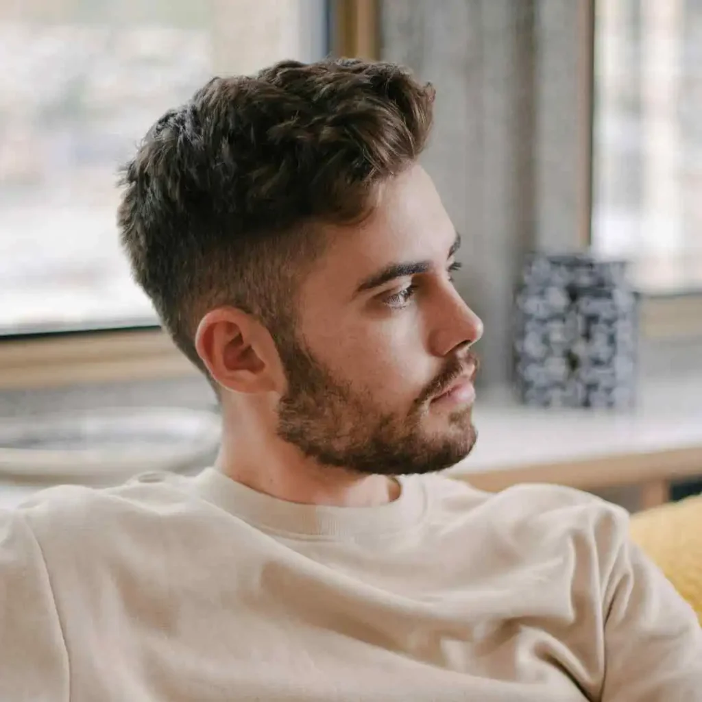 21 On-Trend Hairstyles for Men with Oval Face 3 A young man with short, tousled hair styled for men hairstyles oval face sits casually on a couch, gazing towards a window, with a cozy indoor setting in the background.