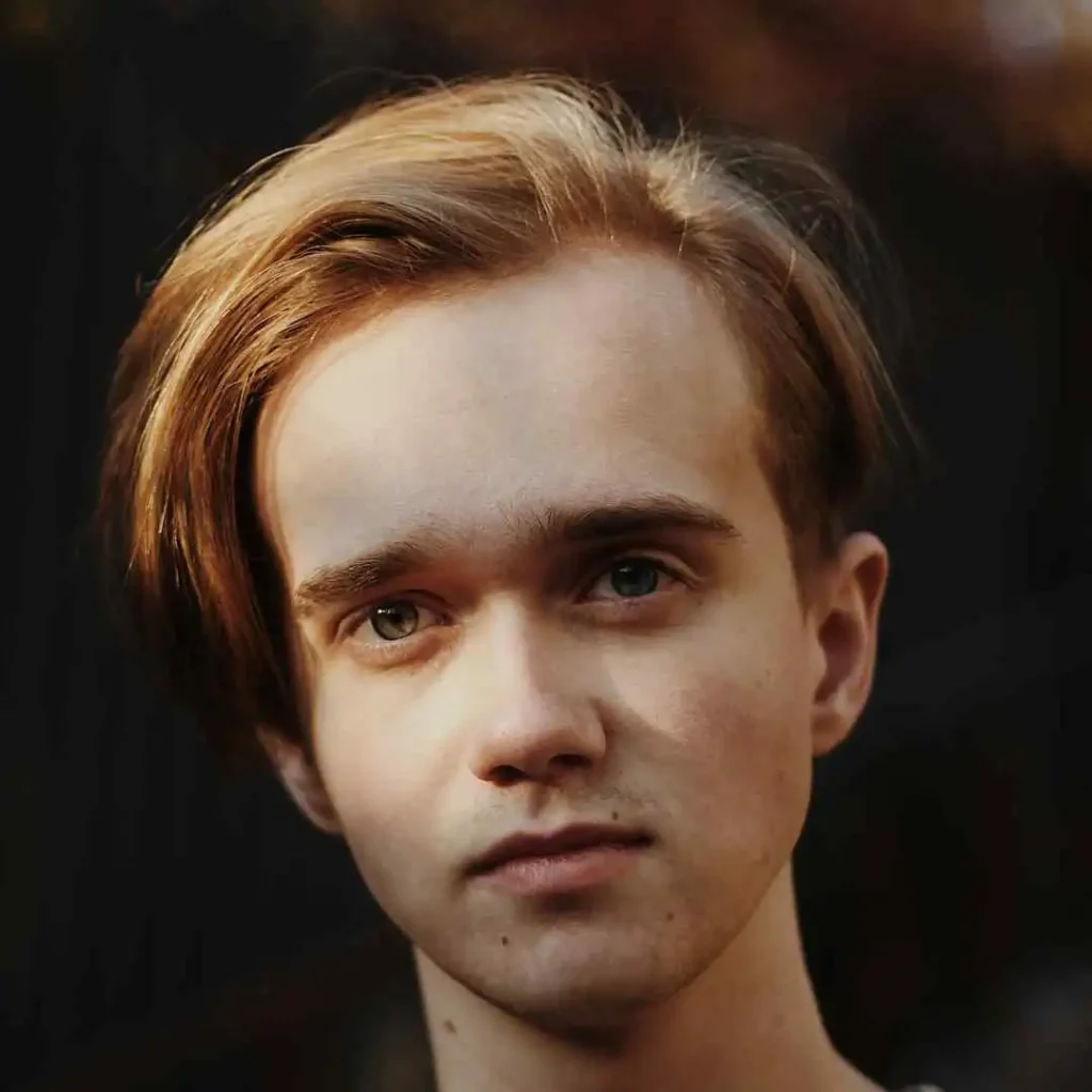 A close-up portrait of a person with light brown hair styled in soft waves, set against a blurred dark background.