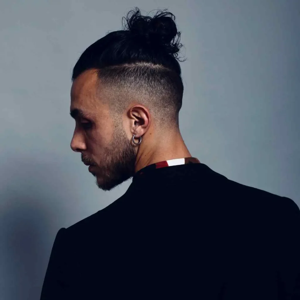 Person with a stylish bun, wearing a black jacket and a colorful collar, poses against a gray background.