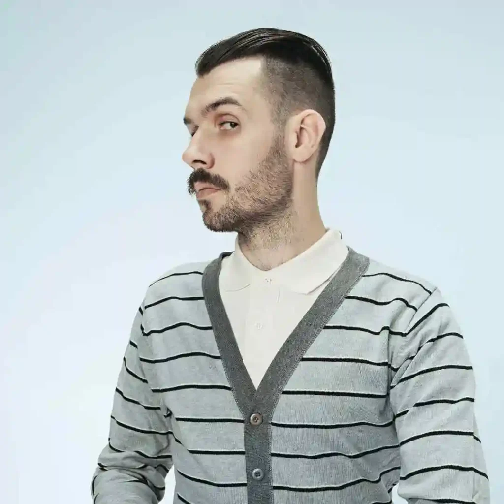 A person wears a gray striped cardigan over a white polo shirt against a light background, posing confidently.