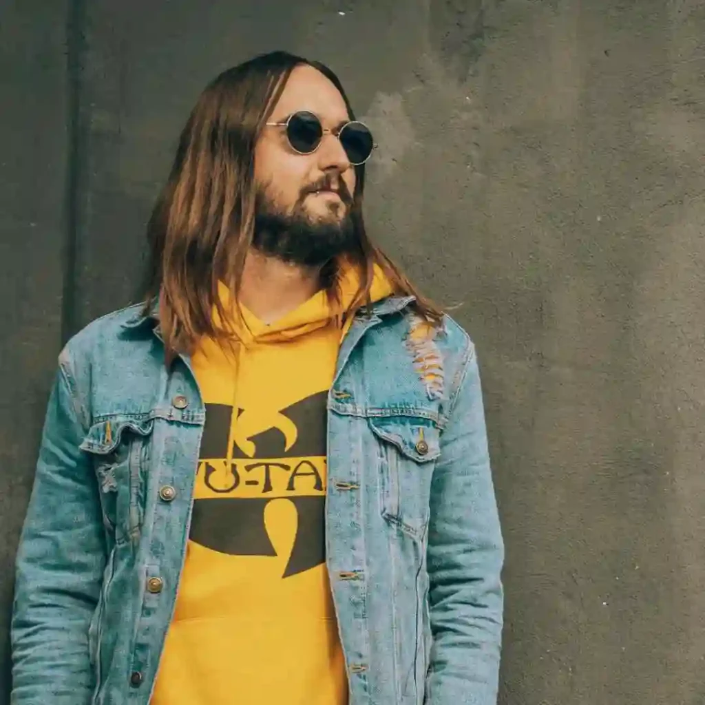 A person stands against a textured wall, wearing a yellow Wu-Tang hoodie layered under a distressed denim jacket.