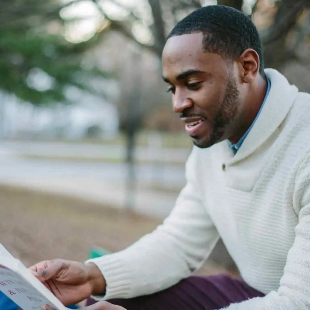 Top 15 Men Hairstyles for Buzz Cut 4 A person with MEN HAIRSTYLES BUZZ CUT, wearing a white sweater, sits outdoors reading a book in a park surrounded by trees.