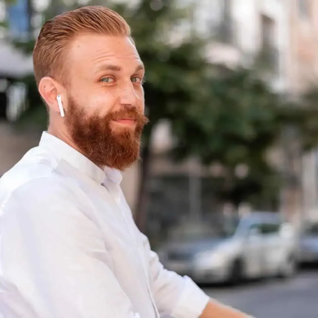 Discover 20 New Pompadour Hairstyles- A Comprehensive Guide 4 A close-up of a man with a red beard and earbuds, wearing a white shirt, standing on a city street with cars in the background, showcasing a stylish pompadour hairstyle.