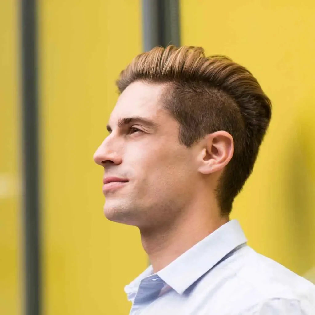 18 Top Men Hairstyles Quiff 4 A profile view of a young man showcasing MEN HAIRSTYLES QUIFF with stylish blonde hair, wearing a light blue shirt against a bright yellow background