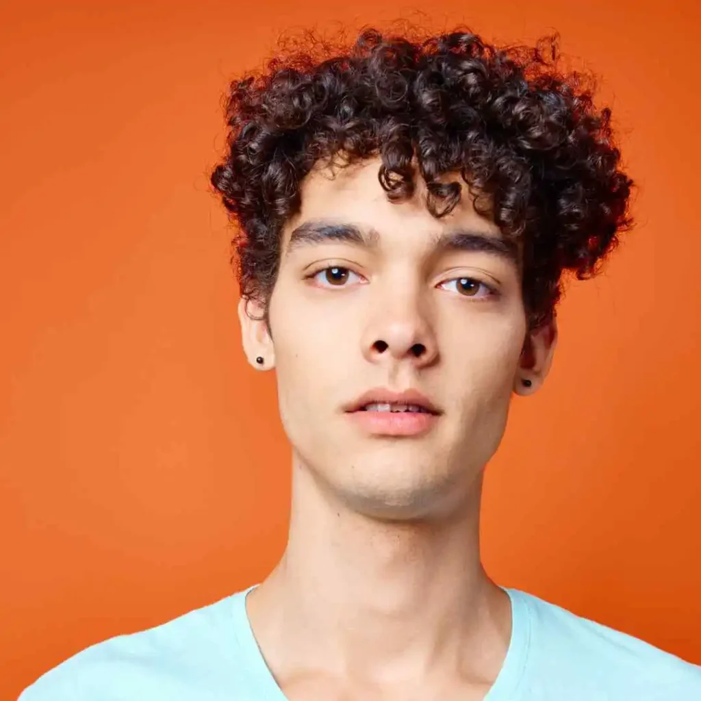 29 Best Men’s Short Curly Haircuts 4 A young person showcasing a trendy men's short curly haircut, wearing a light blue shirt and standing against a bright orange background.