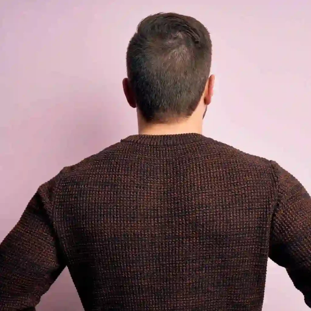 A man with short hair wearing a textured, dark brown sweater stands with his back to the camera against a light pink background.