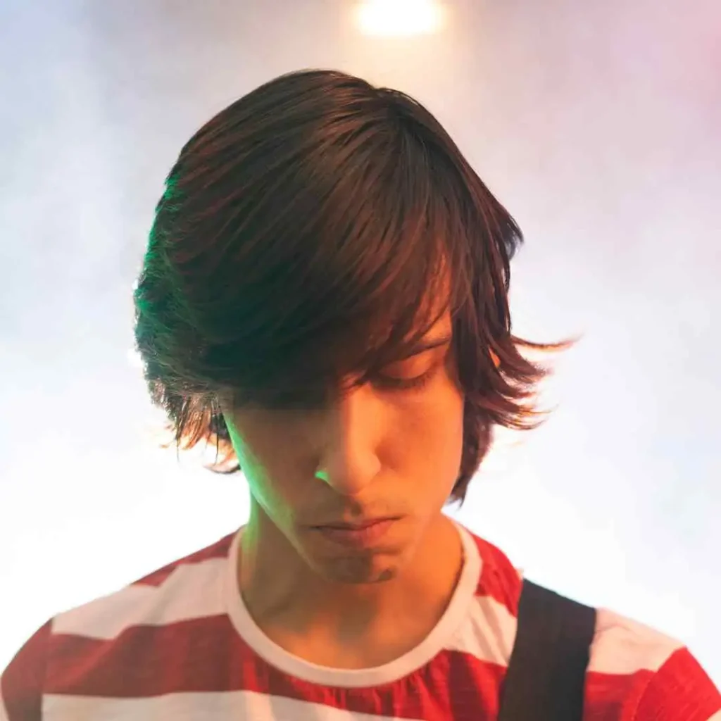 A young person with medium-length hair wearing a red and white striped shirt, standing in front of a colorful, smoky background.