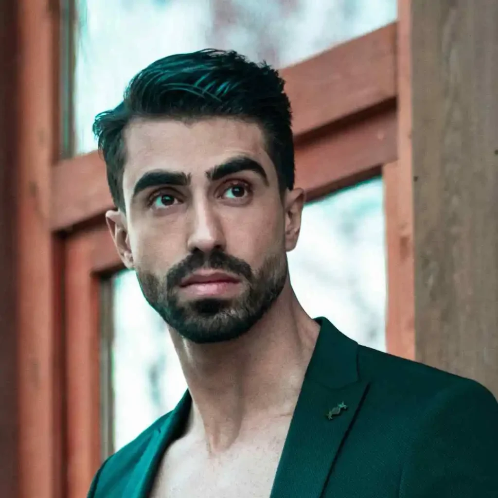 A stylish individual in a green blazer stands against a wooden background, exuding confidence and charm.