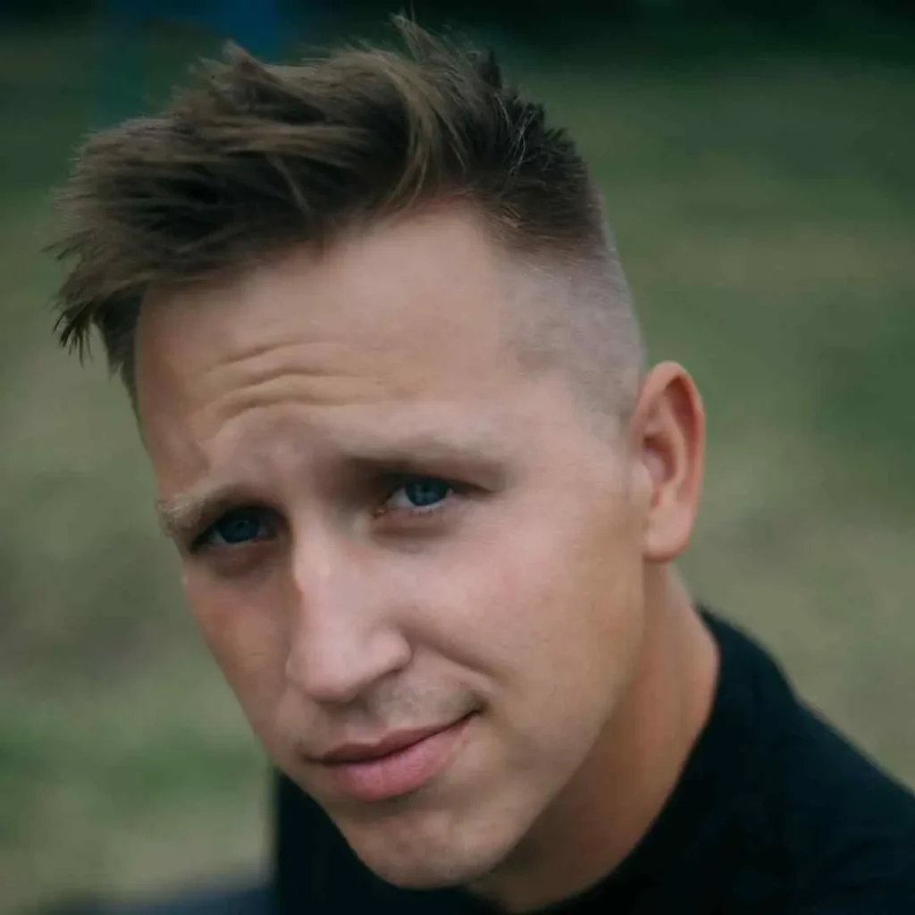 A person with stylish, tousled hair stands outdoors, with a blurred background of green grass and soft natural light.