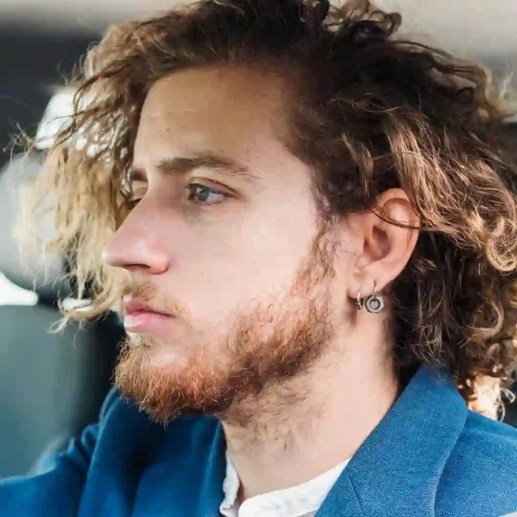 A person with curly hair wearing a blue blazer and a white shirt, seated in a car, focused on the road ahead.