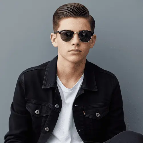 A young man wearing a black denim jacket over a white t-shirt, sitting casually against a light gray background, showcasing one of the trendy teenage guy haircuts.