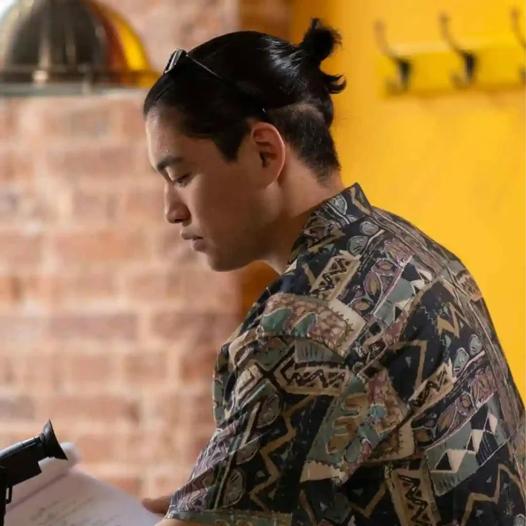 A person in a patterned shirt stands by a black microphone, focused on a document against a brick and yellow background.