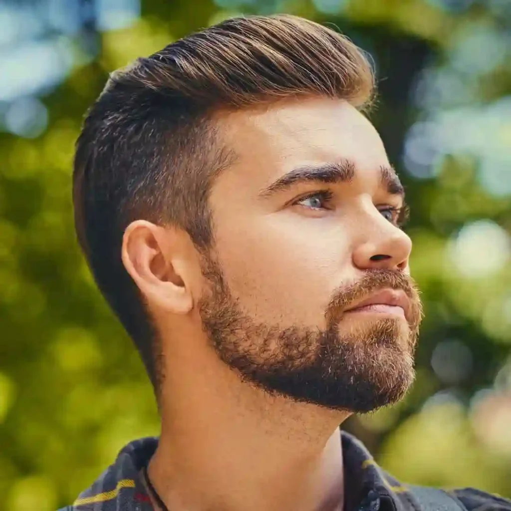 A young man with a stylish haircut stands outdoors, surrounded by greenery, wearing a plaid shirt and a necklace.