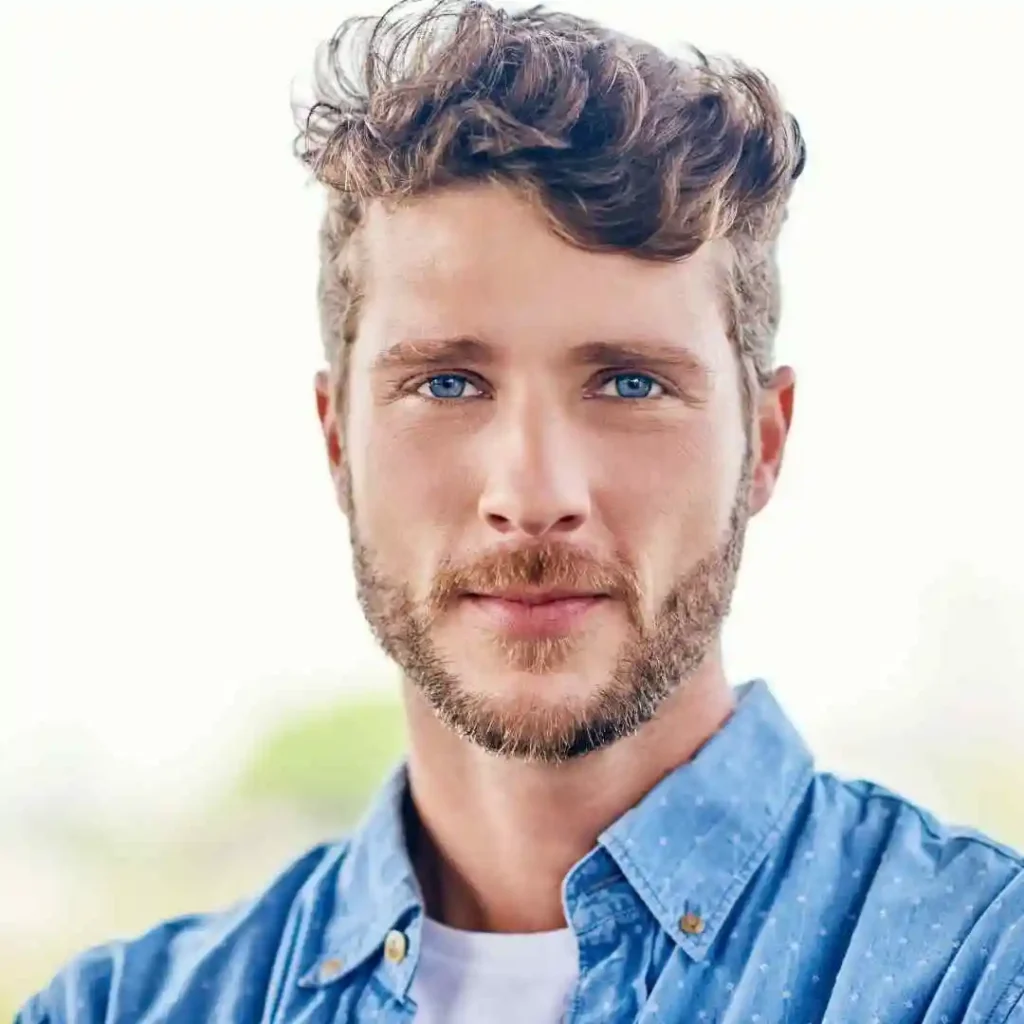 18 Top Men Hairstyles Quiff 5 "A young man with curly hair showcasing MEN HAIRSTYLES QUIFF, wearing a blue button-up shirt over a white t-shirt, standing outdoors with a light background.