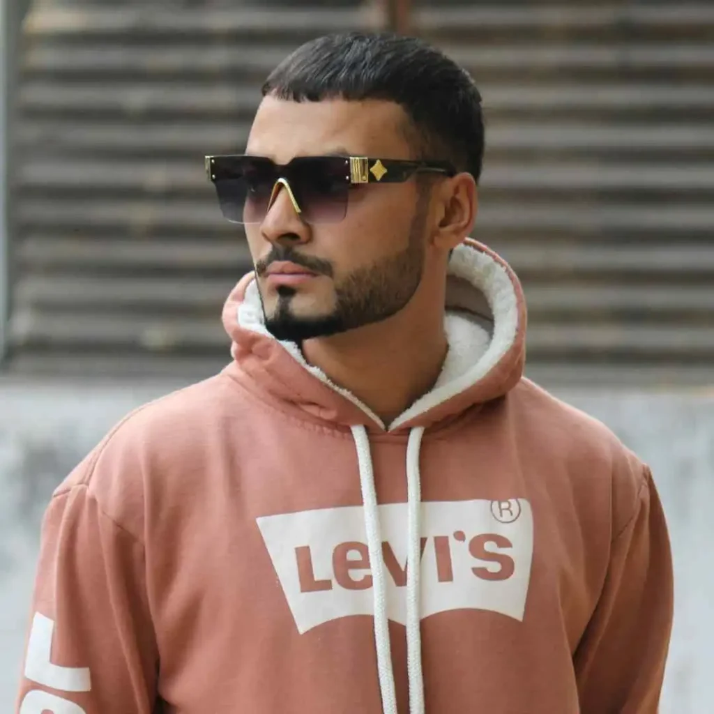 Top 15 Men Hairstyles for Buzz Cut 5 Person showcasing MEN HAIRSTYLES BUZZ CUT, wearing a pink Levi's hoodie with a white logo, styled with sunglasses, in front of a blurred wooden backdrop.