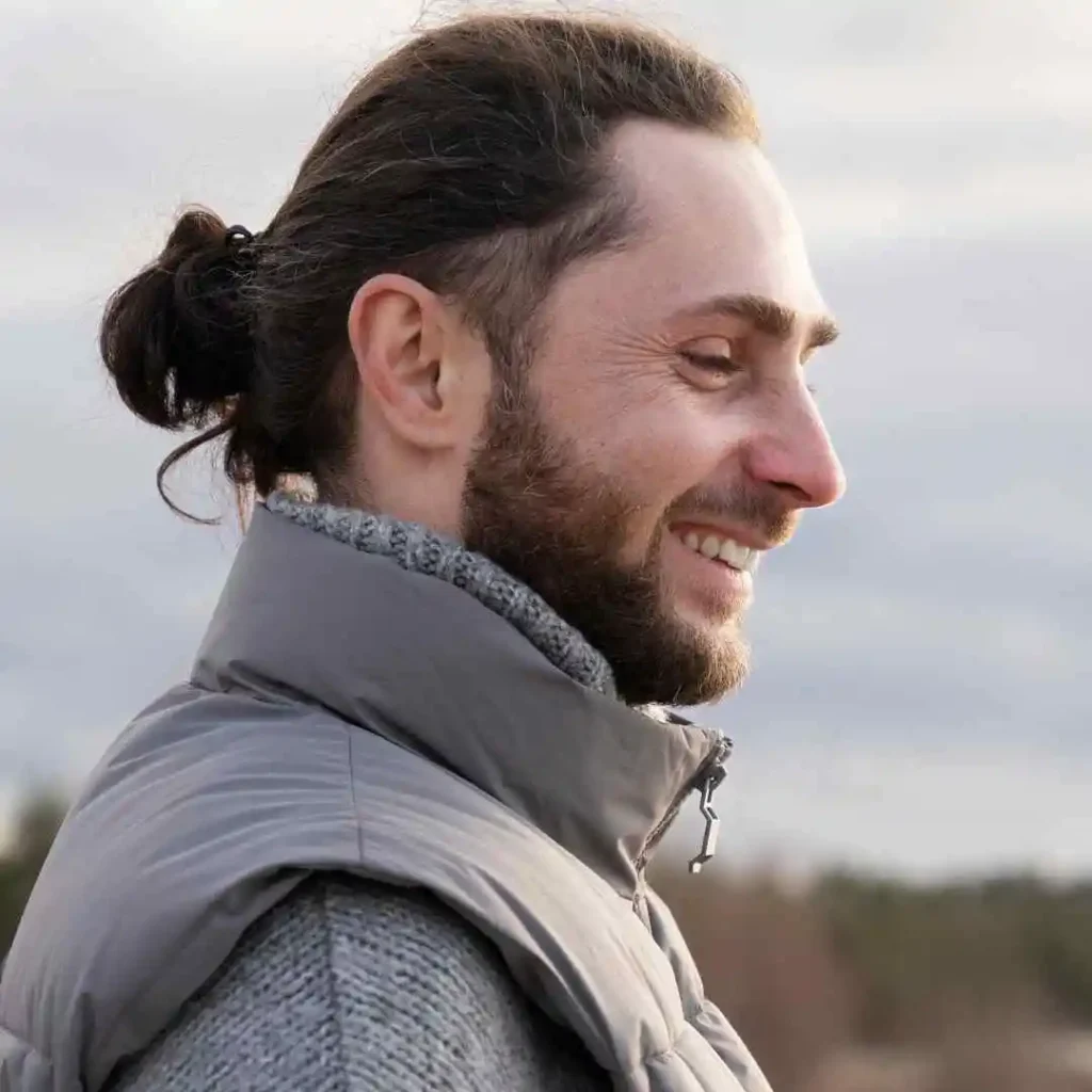 A person with long dark hair tied in a bun, wearing a gray puffy vest over a knitted sweater, stands outdoors against a cloudy sky.