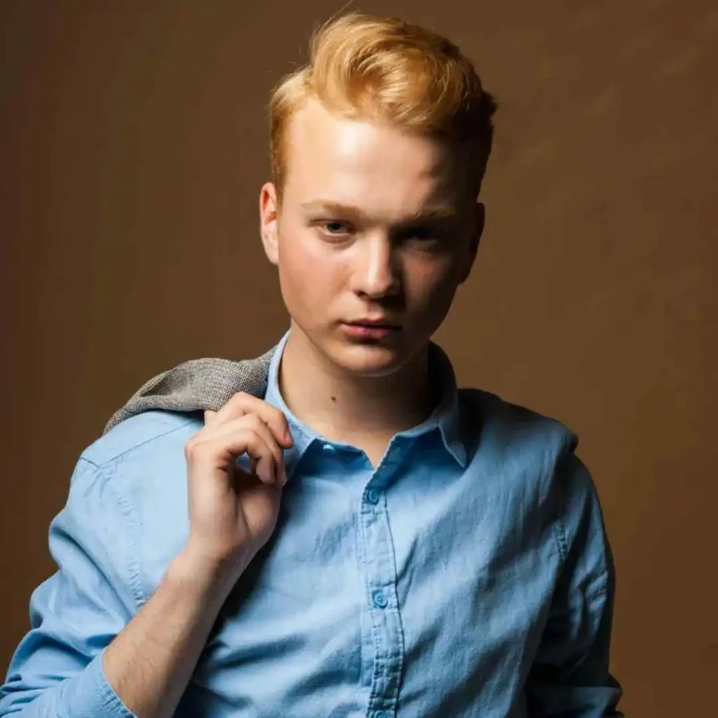 A young man in a light blue shirt holds a grey jacket over his shoulder, standing against a warm brown backdrop.