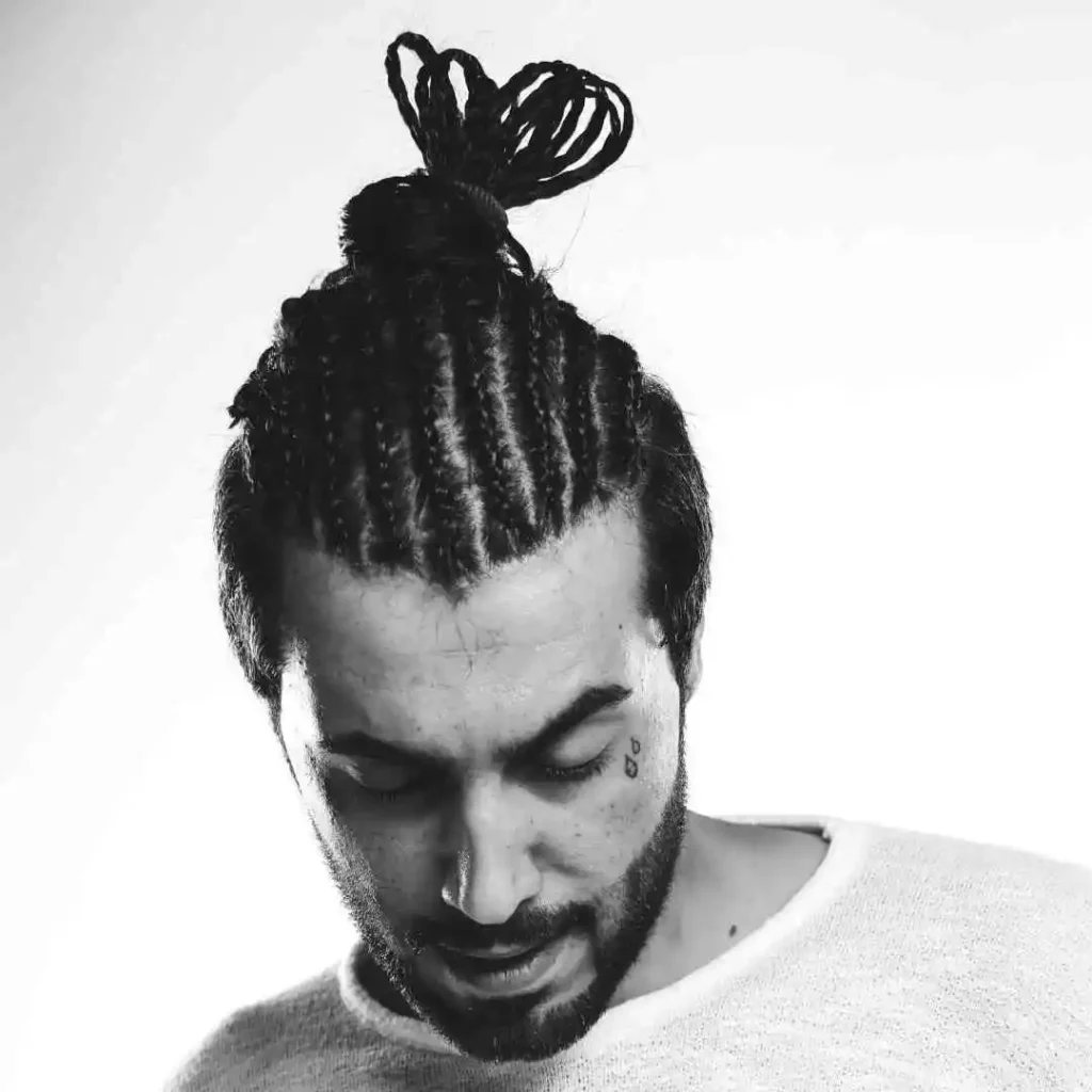A person with braided hair styled in a top knot, wearing a light grey sweater, against a minimalist white background.