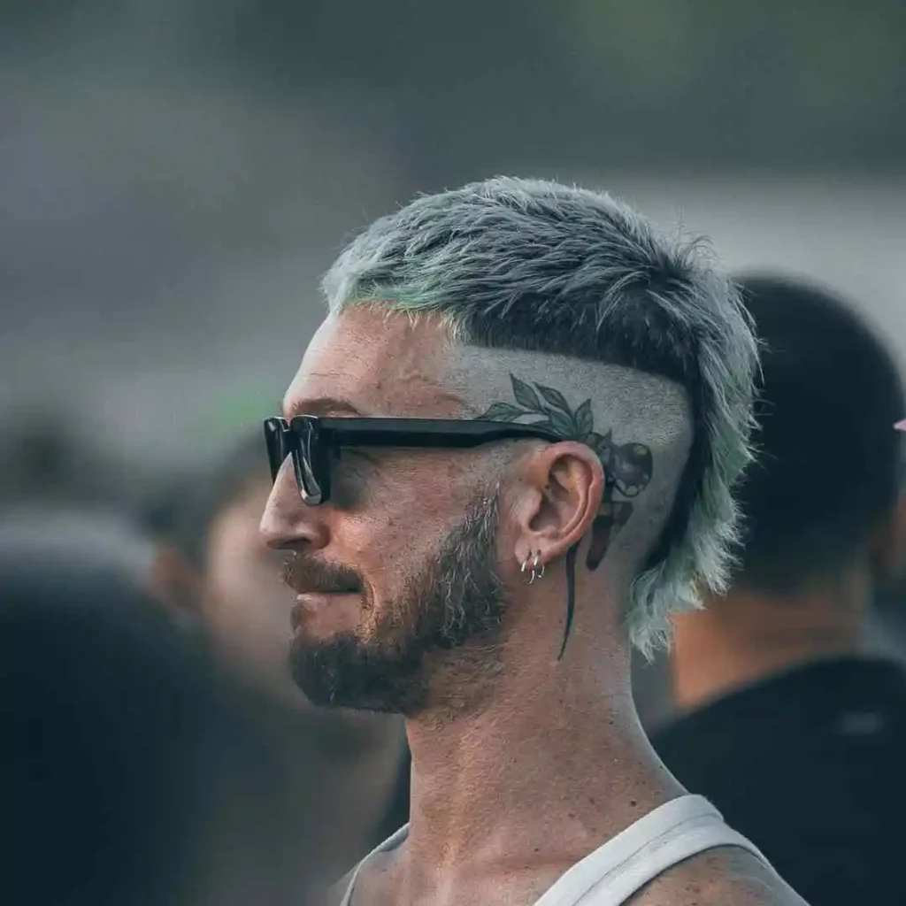 29 Best Men’s Short Curly Haircuts 6 A man with edgy, light blue hair and a tattoo of leaves and fruit on the side of his head, wearing a tank top surrounded by people.
