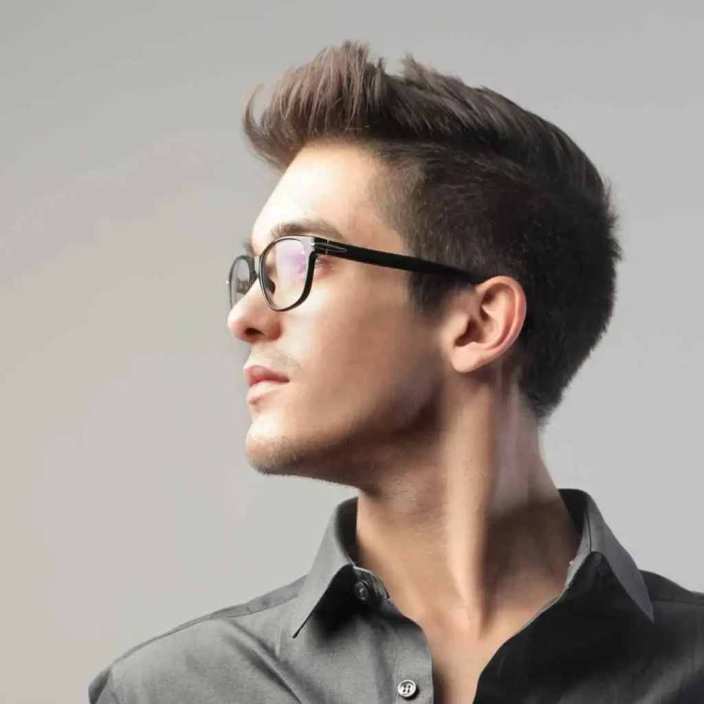 18 Top Men Hairstyles Quiff 6 A man with styled hair featuring a MEN HAIRSTYLES QUIFF, wearing a grey button-up shirt, looks to the side against a neutral background, showcasing a confident posture.
