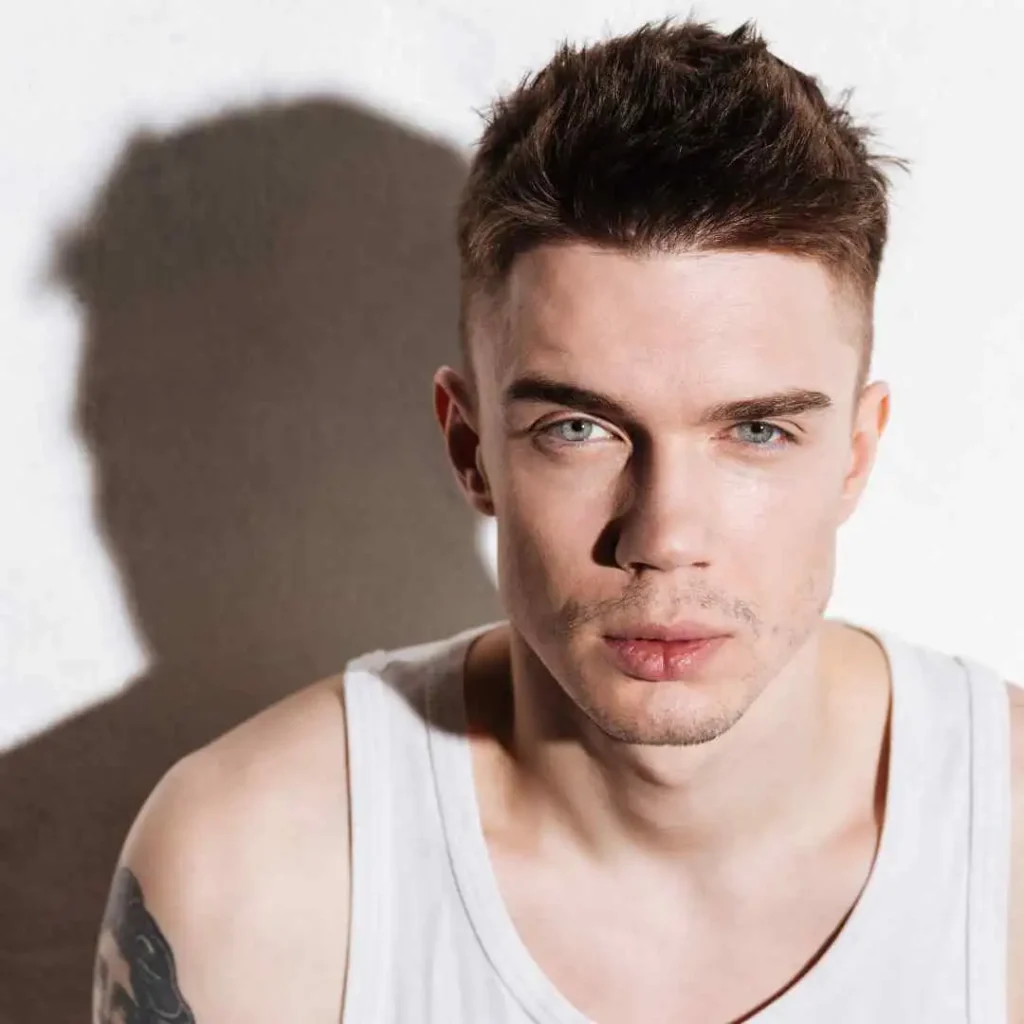Modern Men Short Hairstyles- A Comprehensive Guide 6 A young man with MODERN MEN SHORT HAIRSTYLES in a sleeveless white shirt, with a tattoo on his shoulder, stands against a textured wall, creating soft shadows.