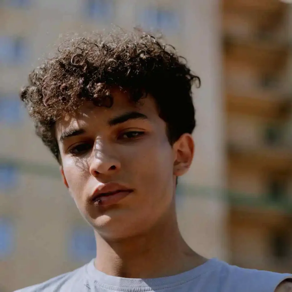 29 Best Men’s Short Curly Haircuts 7 A young person showcasing MEN'S SHORT CURLY HAIRCUTS, wearing a light gray shirt, stands outdoors with a blurred building background.