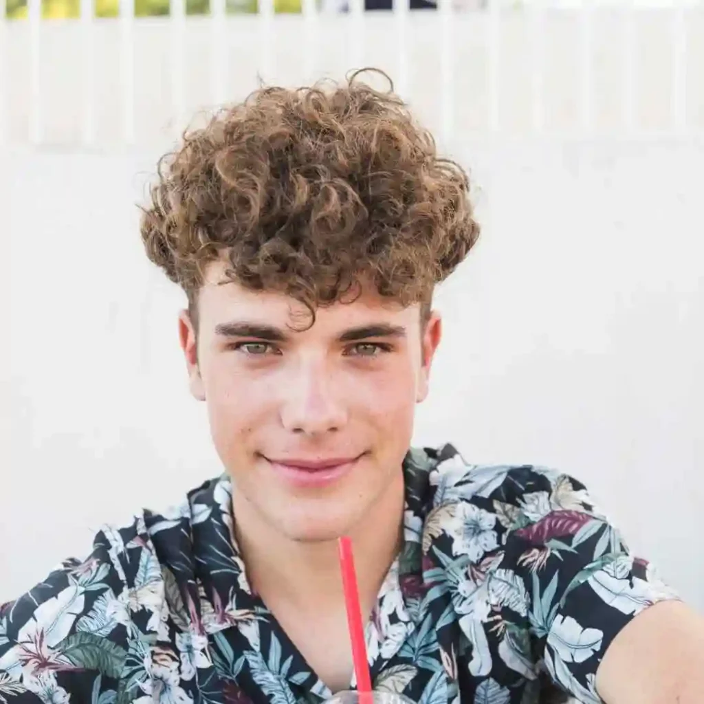 Discover 20 New Pompadour Hairstyles- A Comprehensive Guide 7 A young person with curly hair sporting a pompadour hairstyle is enjoying a drink through a red straw, wearing a vibrant floral-patterned shirt against a white background.