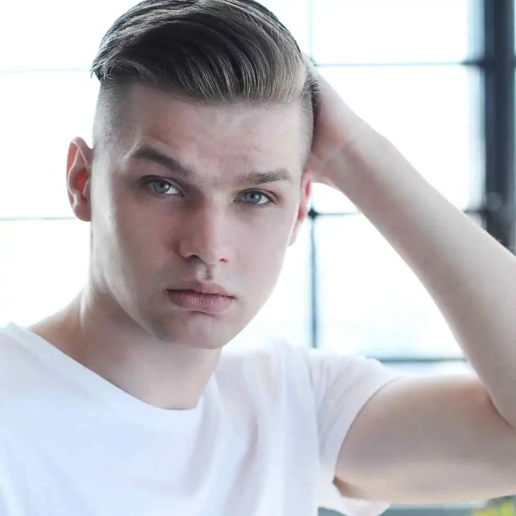Modern Men Short Hairstyles- A Comprehensive Guide 7 A young man in a white t-shirt is posing with one hand behind his head, showcasing a stylish haircut that represents modern men short hairstyles against a bright window backdrop.