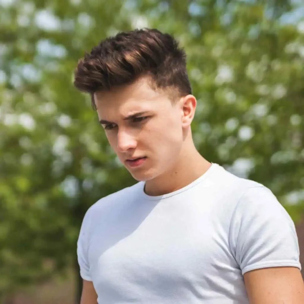 18 Top Men Hairstyles Quiff 7 A young man showcasing MEN HAIRSTYLES QUIFF in a fitted white t-shirt stands outdoors against a backdrop of blurred green foliage.