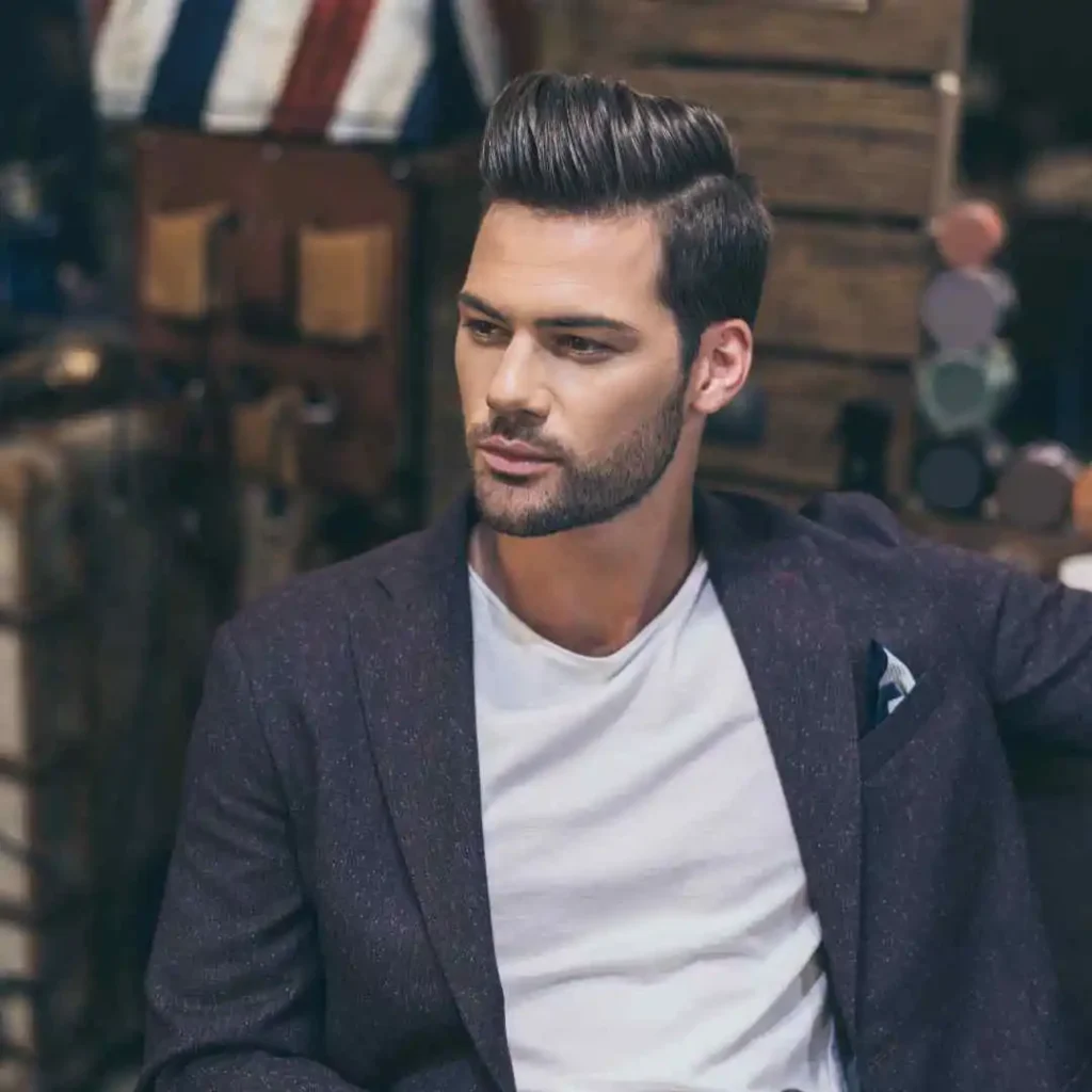 18 Top Men Hairstyles Quiff 8 A stylish man in a dark blazer and white t-shirt sits confidently in a vintage setting, surrounded by old tools and decor, showcasing a sharp men hairstyles quiff.