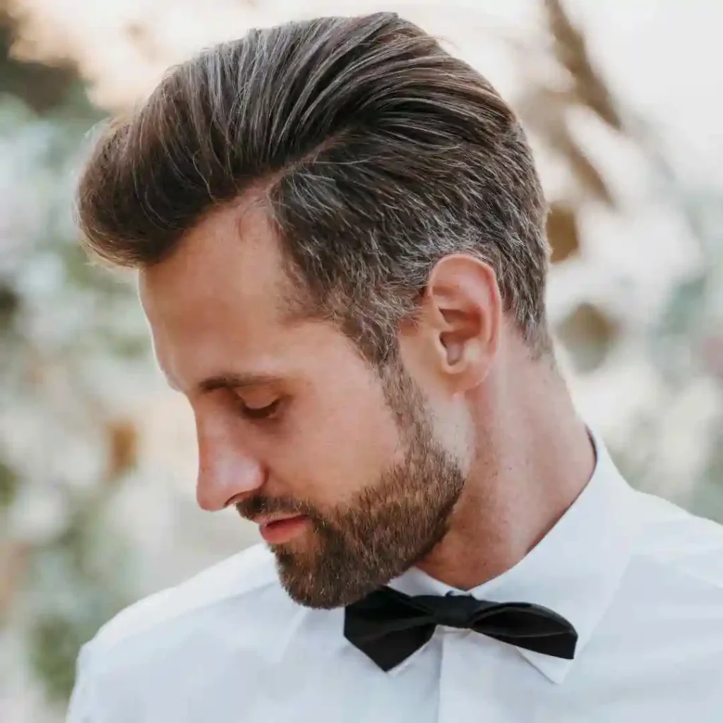 Discover 20 New Pompadour Hairstyles- A Comprehensive Guide 8 A man in a formal white shirt and black bow tie stands outdoors with greenery blurred in the background. His hair is styled neatly in a pompadour hairstyle.