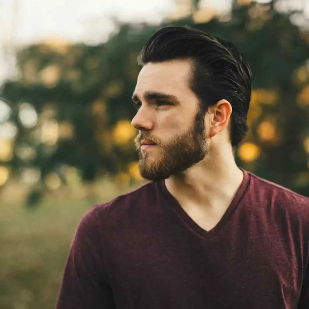 A man in a maroon V-neck shirt showcases MODERN MEN SHORT HAIRSTYLES outdoors, with a softly blurred background of greenery and warm, golden light.