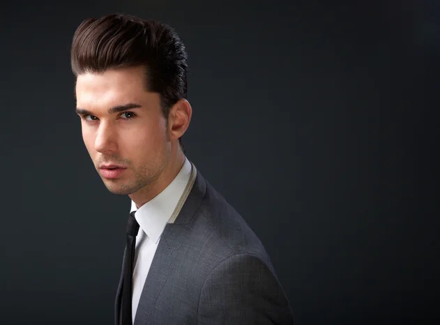 "A man with POMPADOUR HAIRSTYLES in a tailored gray suit with a white shirt and black tie poses against a dark background, showcasing a stylish and modern look.