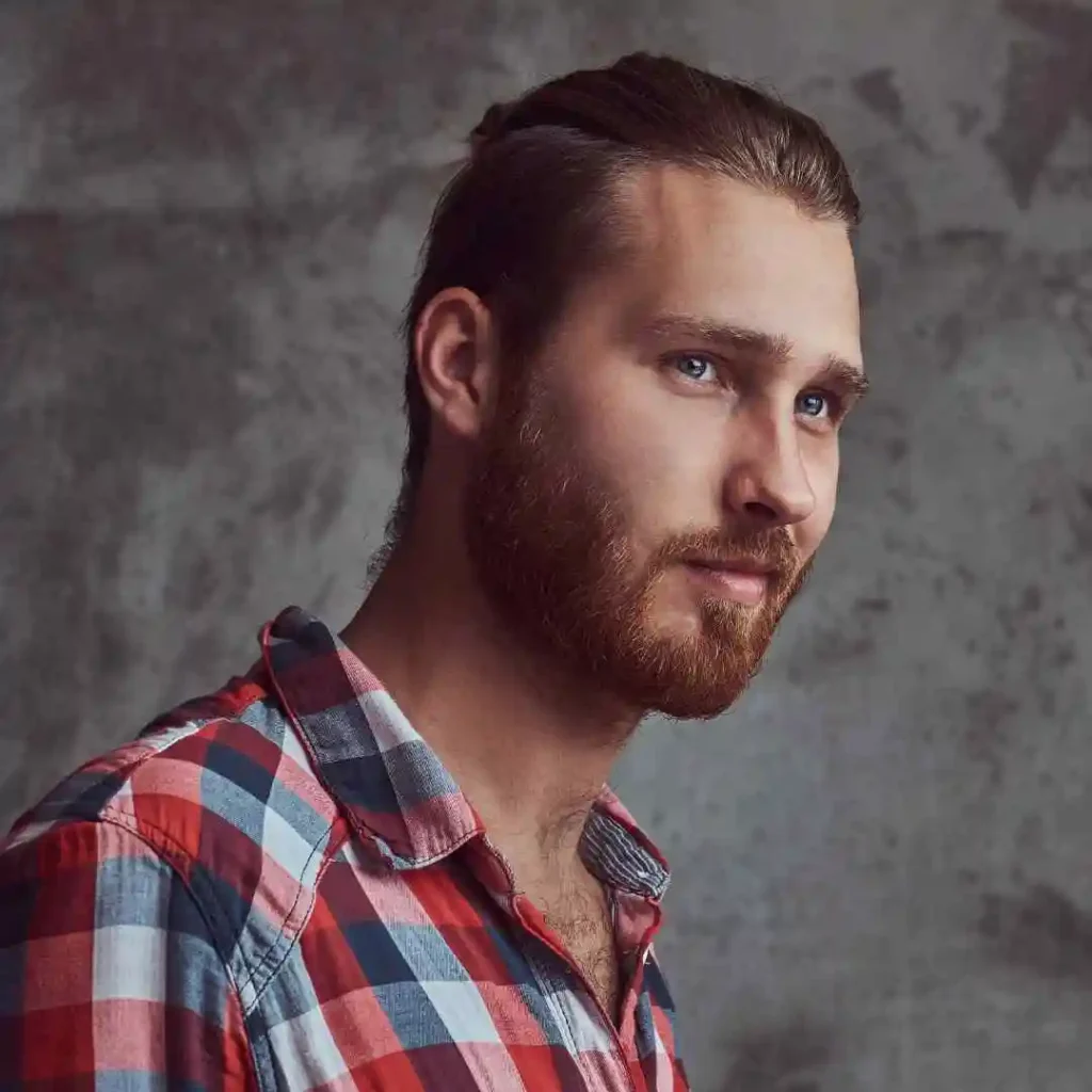 A man in a red and blue checkered shirt stands against a textured grey wall, showcasing his side profile, casual style, and a clean MEN HAIRSTYLES BUZZ CUT.