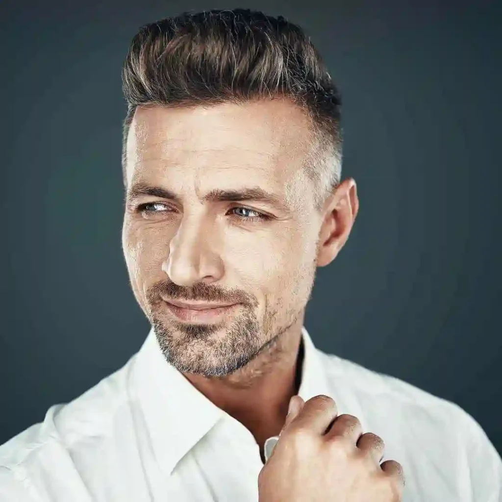 A man showcasing MEN HAIRSTYLES QUIFF in a white shirt poses confidently against a dark gray background, lightly touching the collar of his shirt.