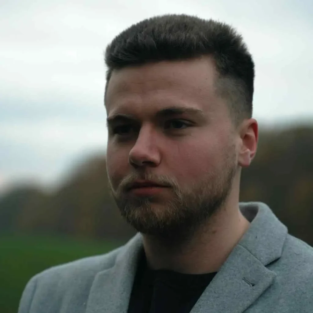 A person showcasing Men Hairstyles for Oval Face, wearing a gray blazer outdoors, with a blurred background of greenery and cloudy skies