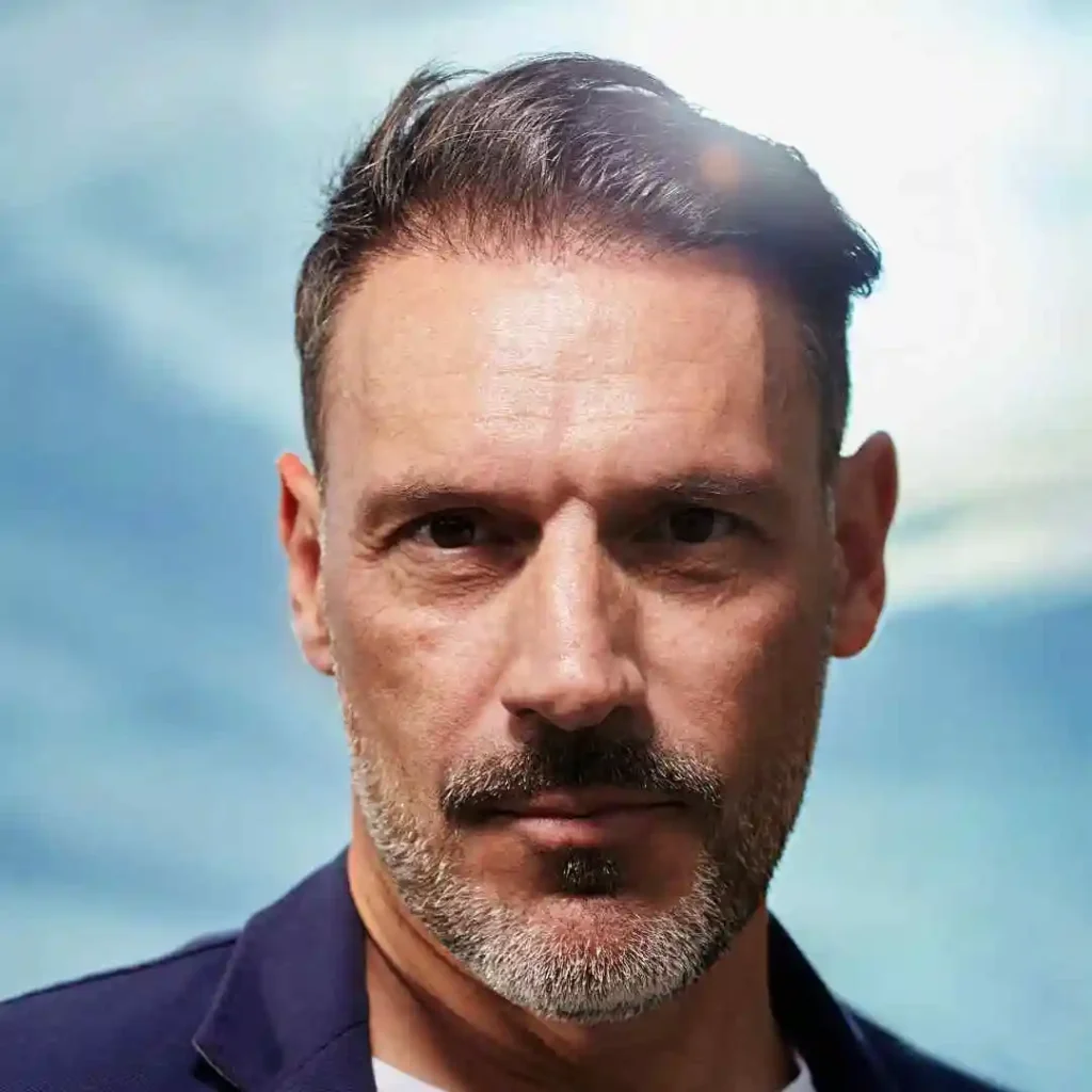 A man with dark, styled hair stands against a bright blue sky, wearing a navy blazer and white shirt, enjoying the sunlight, showcasing an Edgar haircut for men.
