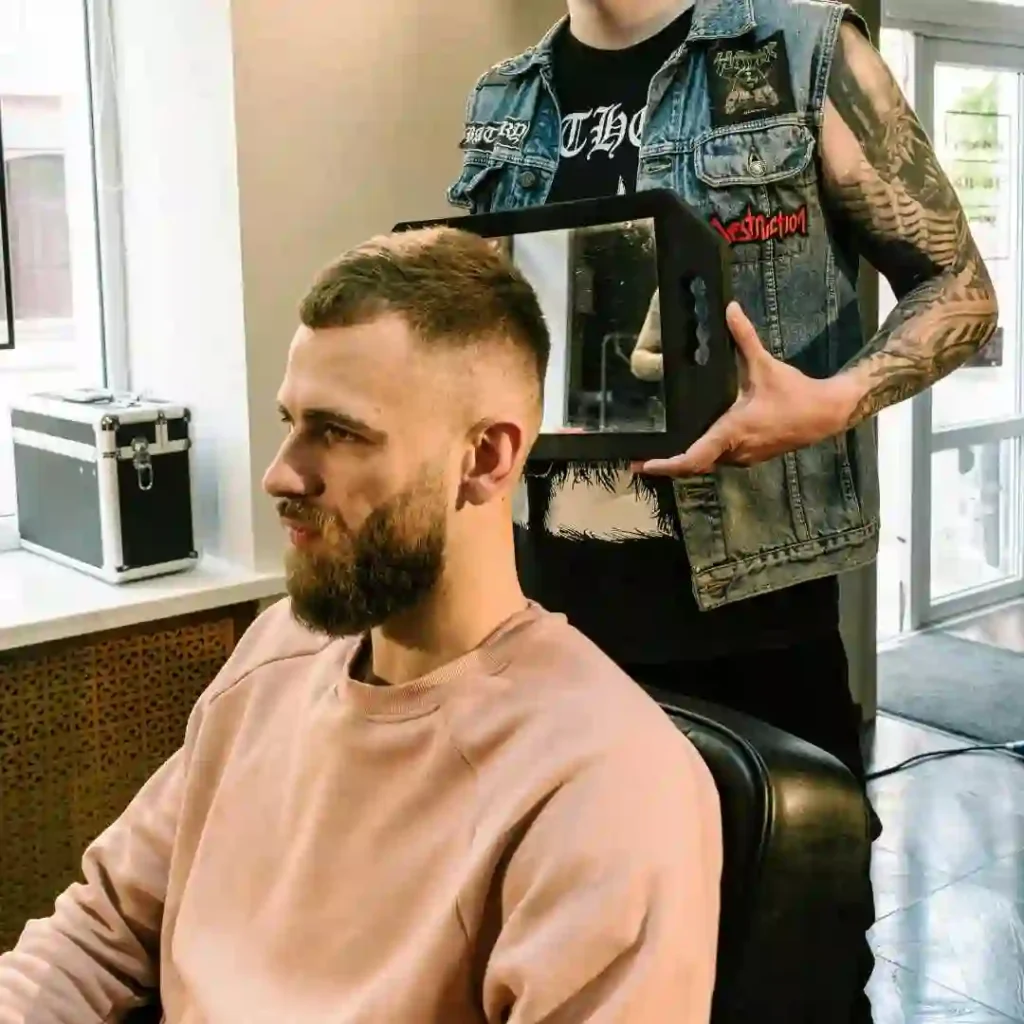 A person sits in a salon chair while a barber holds a mirror behind their head, showcasing a fresh Edgar haircut for men.