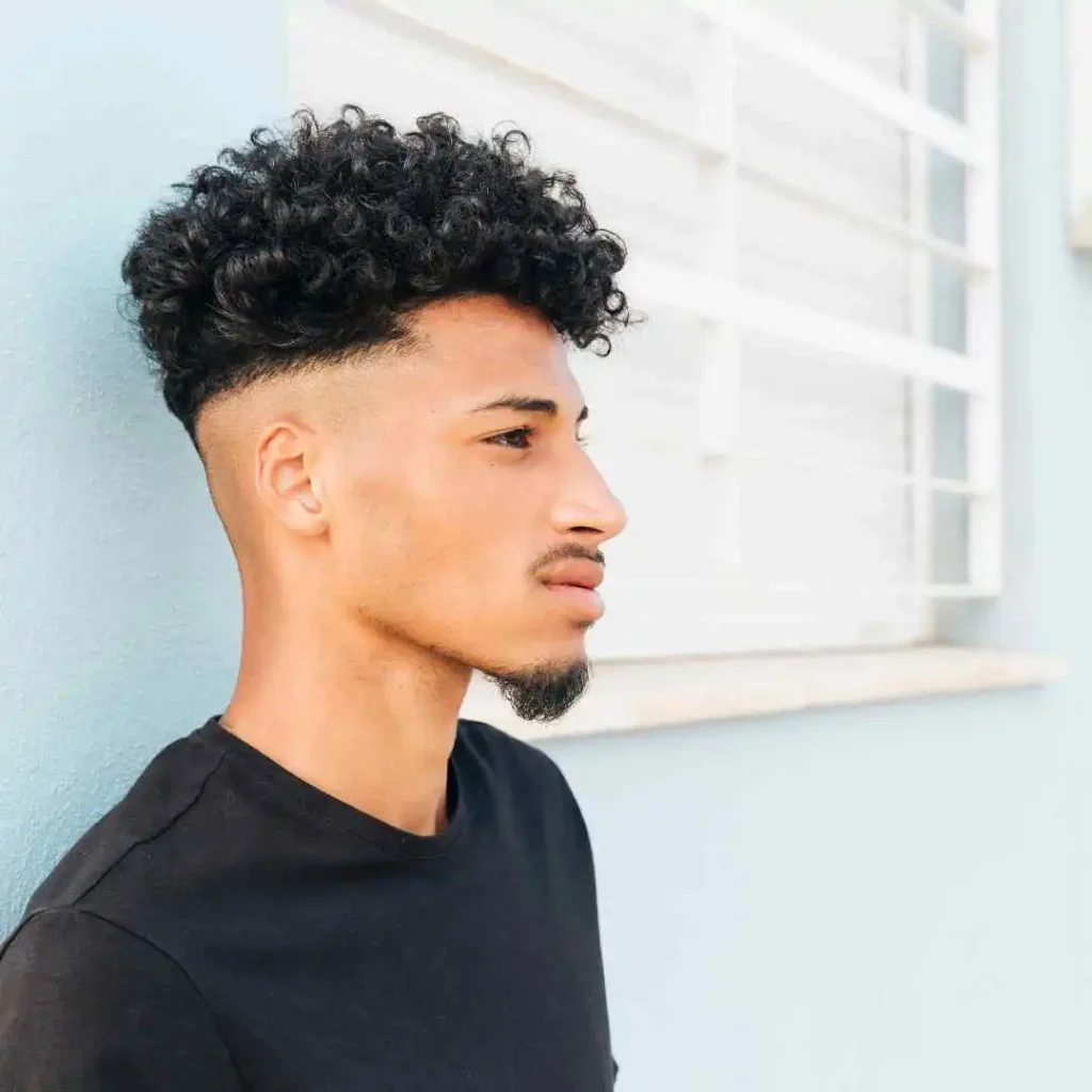 Edgar Hairstyles for Men- Top Styles You Needs to Try 4 A person with a stylish curly top hairstyle, showcasing Edgar haircuts for men, stands against a light blue wall wearing a black t-shirt. Sunlight highlights the scene.