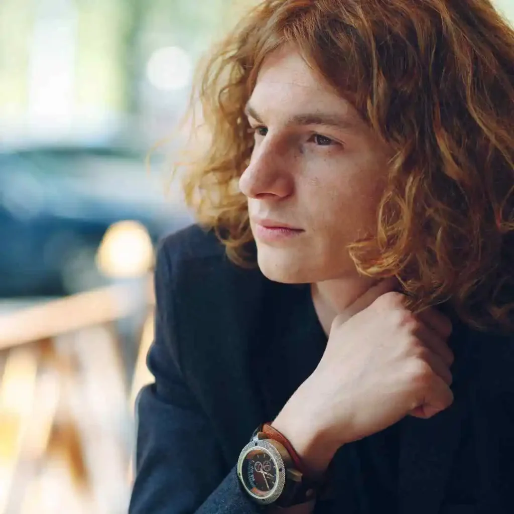 A close-up of a person with curly red hair resting their chin on their hand, wearing a stylish wristwatch, showcasing men hairstyles curtains, against a blurred background.