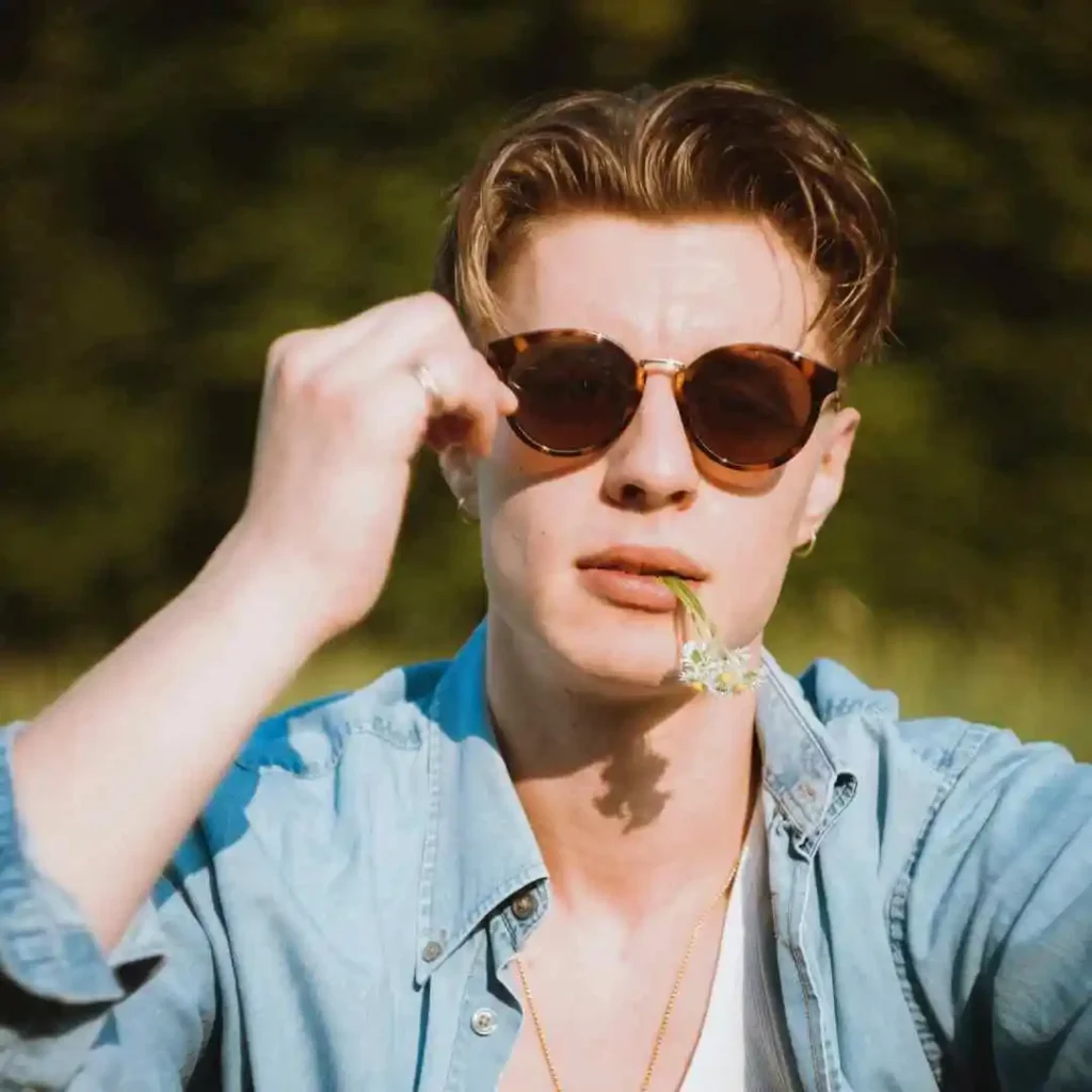 A young man in a denim shirt poses outdoors, holding his hand to his hair styled in MEN HAIRSTYLES CURTAINS, with a sunny, green background.