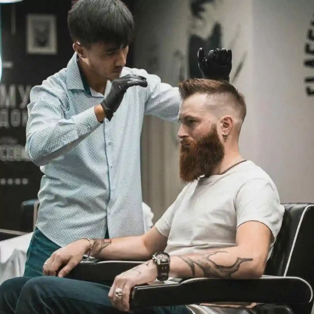 A barber styles a client's hair in a modern salon, showcasing a trendy MEN HAIRSTYLES CURTAINS haircut and visible tattoos on the client's arm.