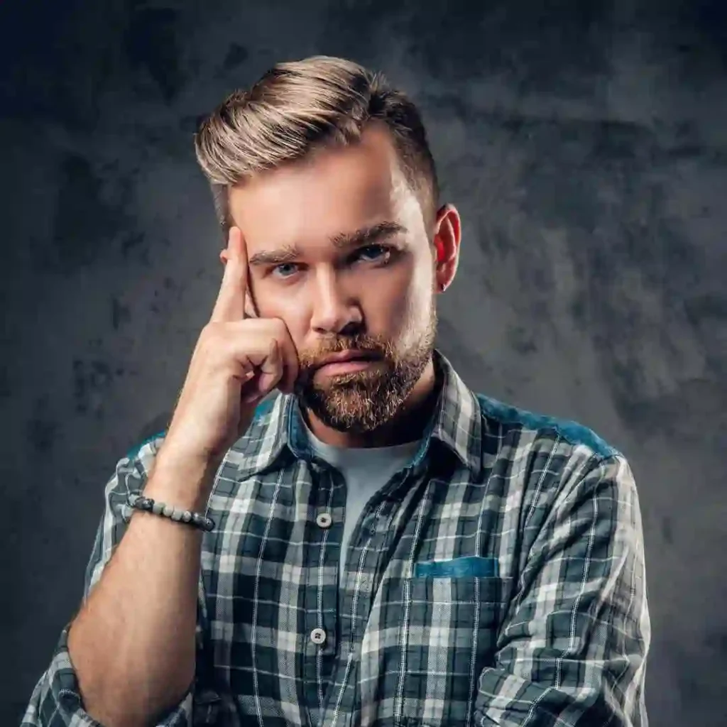 A man in a plaid shirt showcasing Edgar haircuts for men poses thoughtfully against a textured dark background, resting his chin on his hand.
