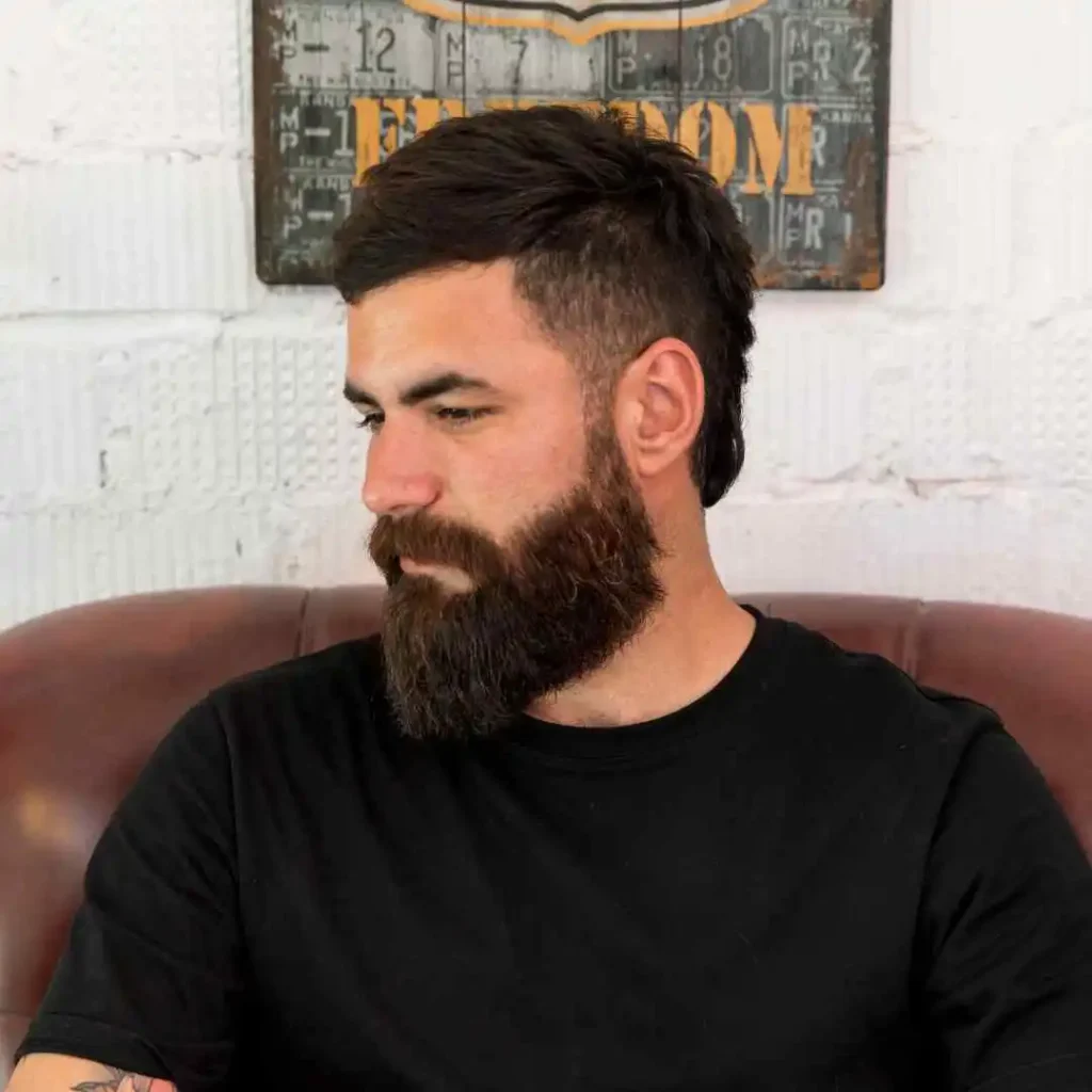 Edgar Hairstyles for Men- Top Styles You Needs to Try 1 A man with an Edgar haircut for men, short dark hair, and a full beard sits casually on a leather sofa, wearing a black T-shirt, in front of a decorative wall.